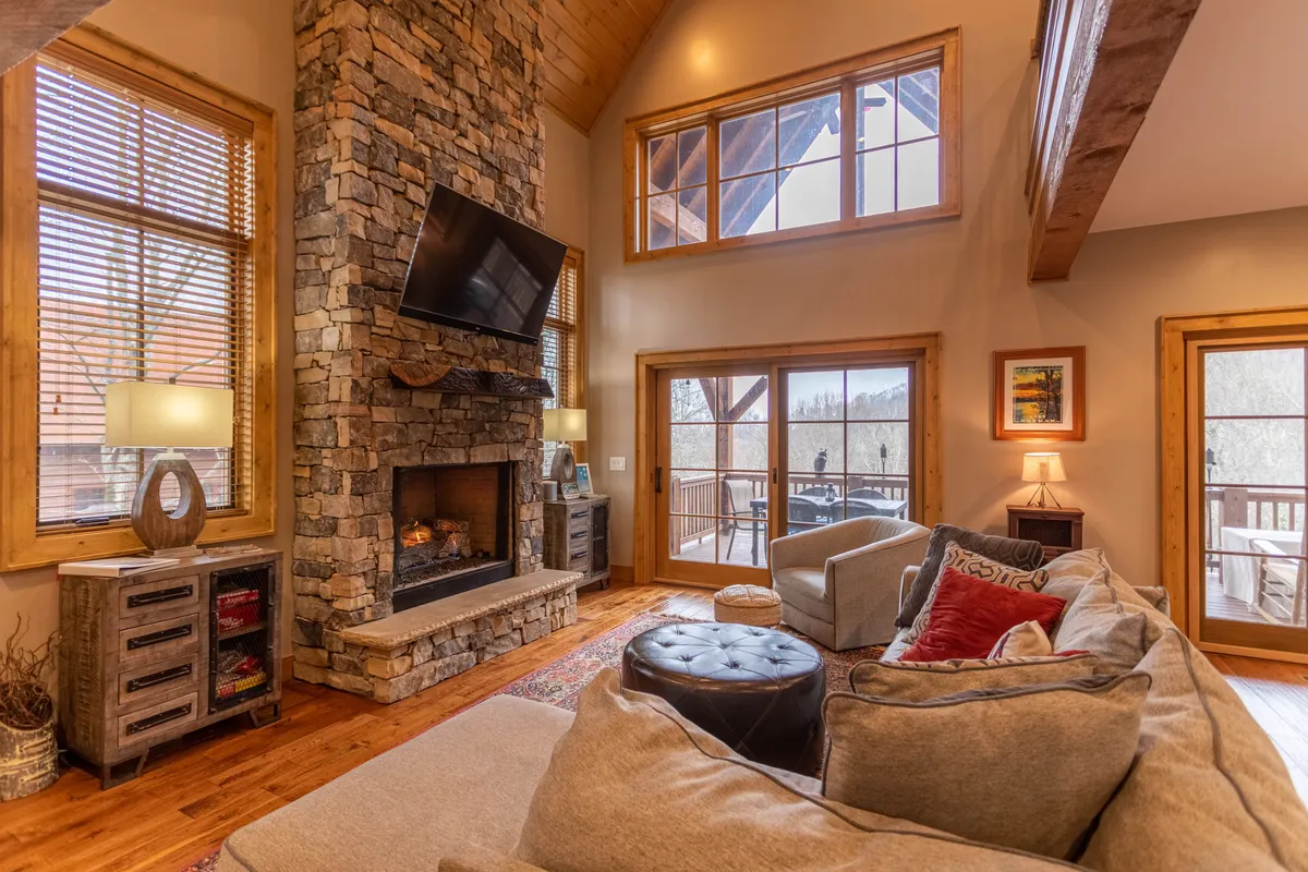 The living room features a comfortable sofa set in front of a stone fireplace, large windows framing outdoor views, and a wooden coffee table. The artwork and decor provide warmth and character, while the windows draw the eye toward sunsets and mountain vistas.
