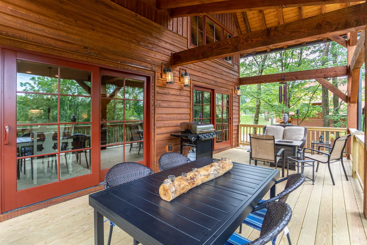 An elevated wooden deck is outfitted with a large outdoor dining table, grill, and a relaxed seating area. Floor-to-ceiling windows reflect the surrounding nature, reinforcing the indoor-outdoor connection, while the space is clearly designed for gatherings and al fresco meals.