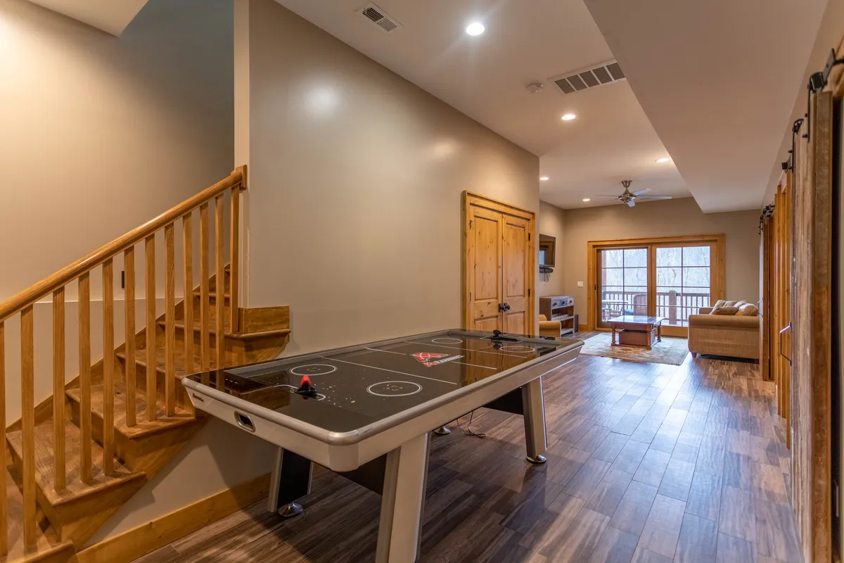 This image showcases a playful lower-level game room featuring a prominent air hockey table, a wooden staircase leading to the upper level, large windows letting in natural light, and a comfortable seating area. The overall ambiance is energetic yet relaxed, with a focus on fun and leisure.