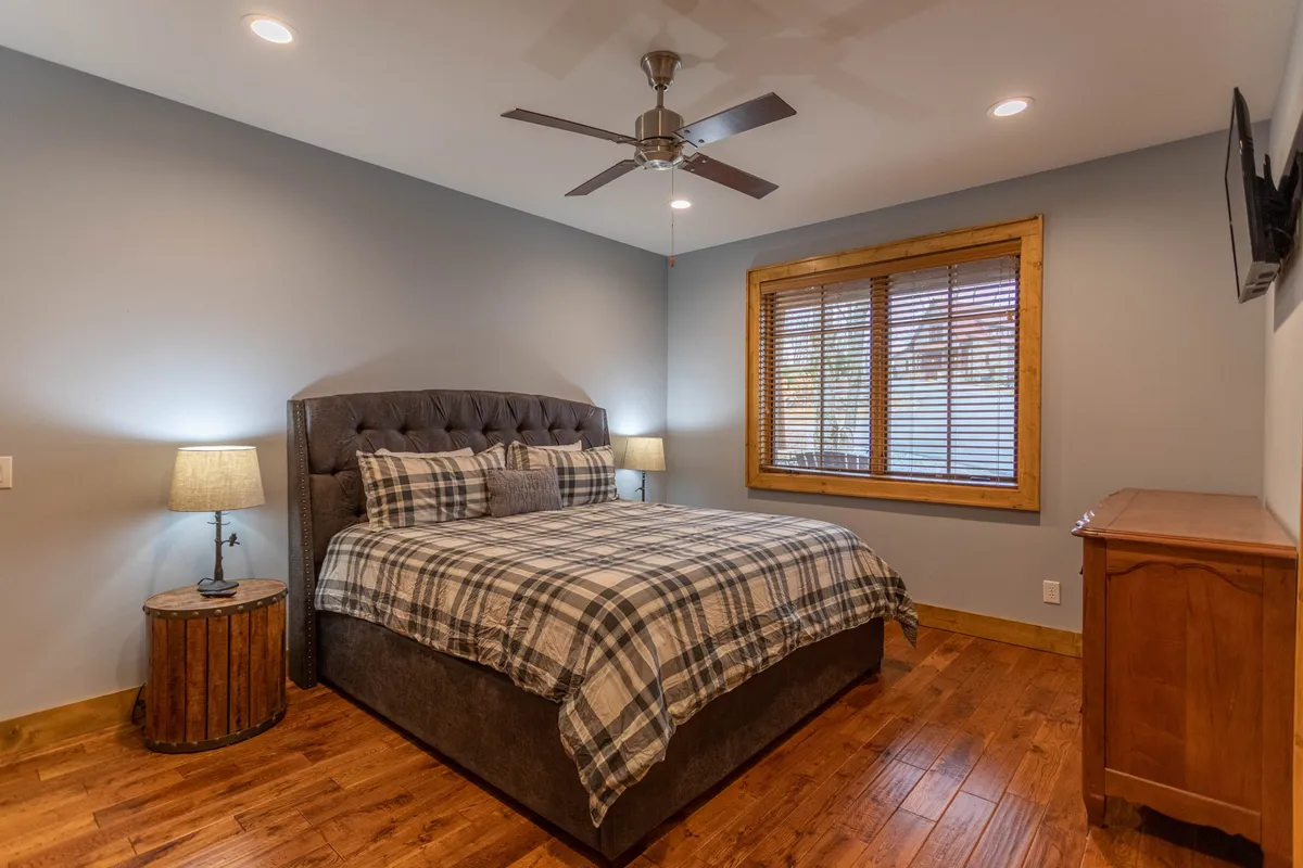 A king bedroom featuring inviting plaid bedding, a tufted headboard, warm wooden furniture, a cozy lamp, and a window with blinds letting in soft natural light. The overall atmosphere is one of warmth and comfort, ideal for restful nights or leisurely mornings.