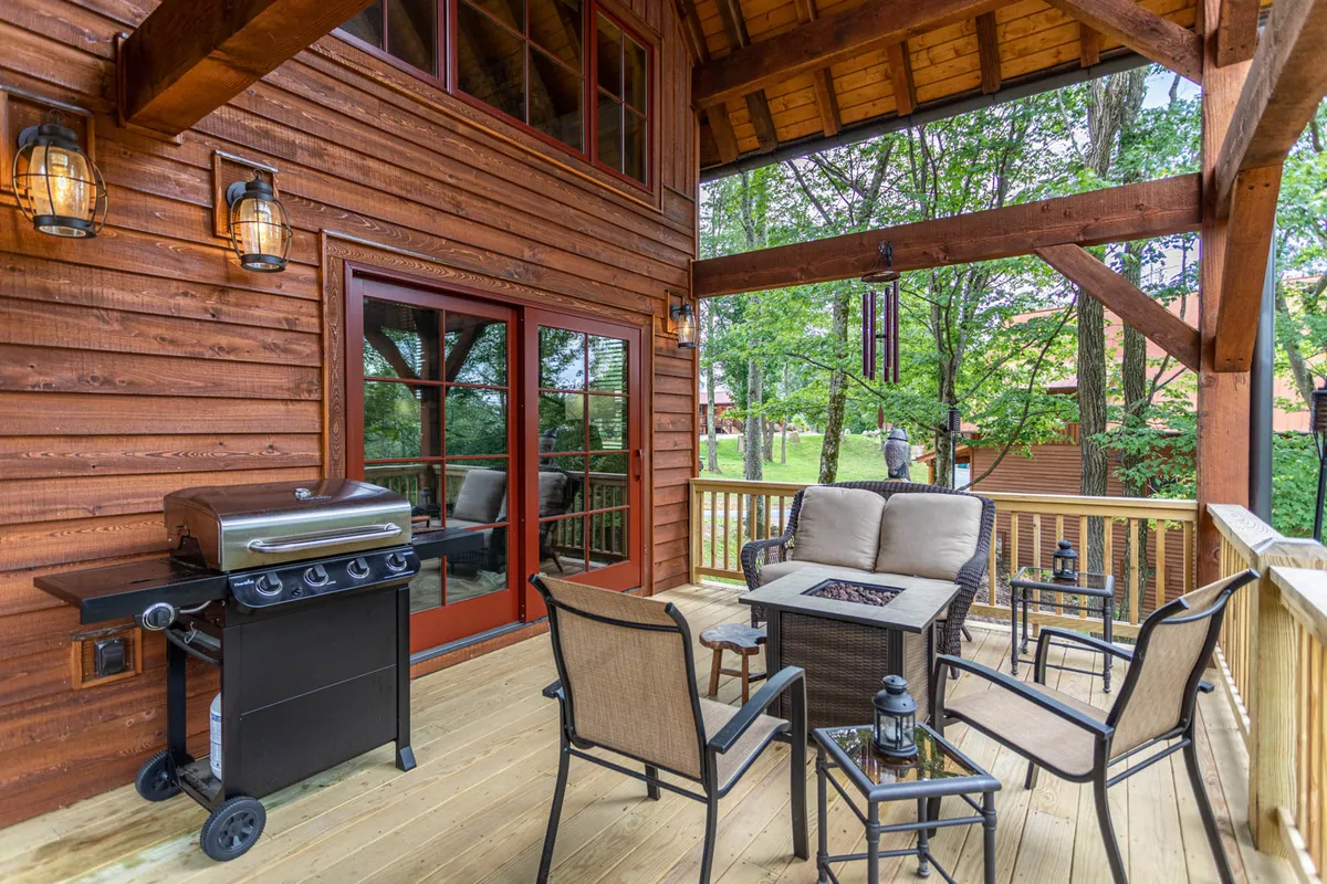 A comfortable outdoor scene featuring a grill, cozy seating area, and a prominent fire pit surrounded by towering trees. The inviting setup suggests both daytime relaxation and evening gatherings around the fire.