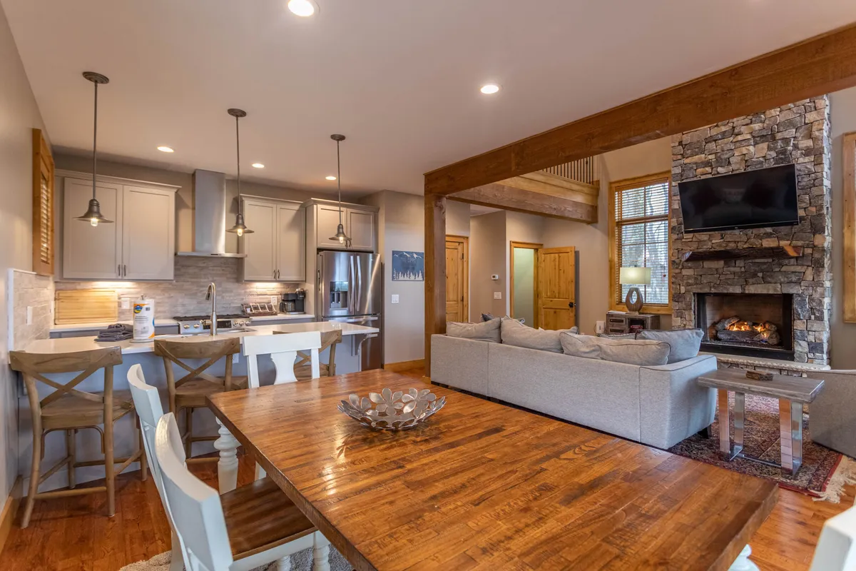 A view of the open-concept great room featuring a dining table set for eight, plush sofa seating, a stone fireplace, and the kitchen island in the background. Natural light floods the space, highlighting both the communal area and the seamless flow between living, dining, and cooking zones.