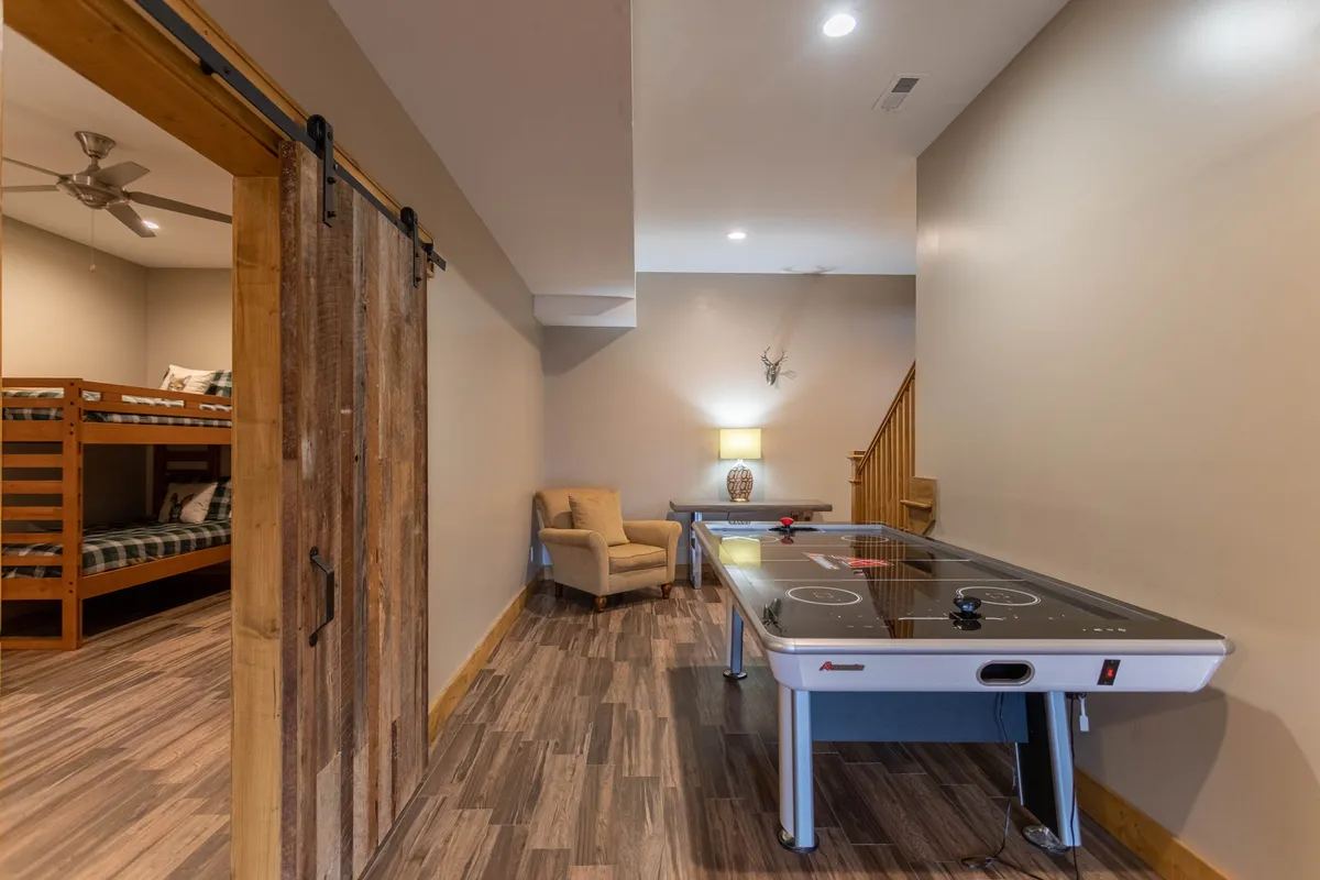 This cozy interior highlights another angle of the game room, featuring an air hockey table, an inviting armchair for spectators, a rustic wooden sliding door, and charming bunk beds in the background. The space feels playful yet homey, ideal for both active play and quiet downtime.