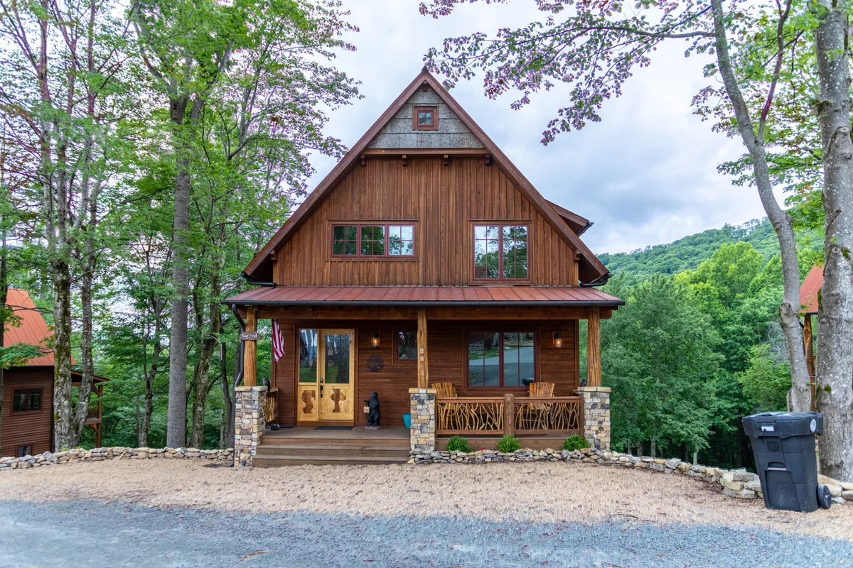 A welcoming view of the cabin's wooden facade, accented with stone, large windows, and a front porch with seating tucked among mature trees. The inviting porch and natural setting emphasize the seamless connection between the home and the outdoors, ideal for relaxing after a day of activities or gathering with family under the stars.