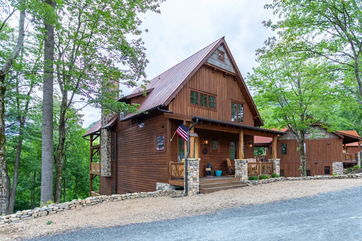 The hero image showcases the home's warm wooden exterior with charming stone accents, a welcoming front porch adorned with a flag, and the inviting outdoor spaces surrounded by mountain scenery. The cozy ambiance hints at opportunities for family gatherings, relaxing evenings, and nature-filled adventures.