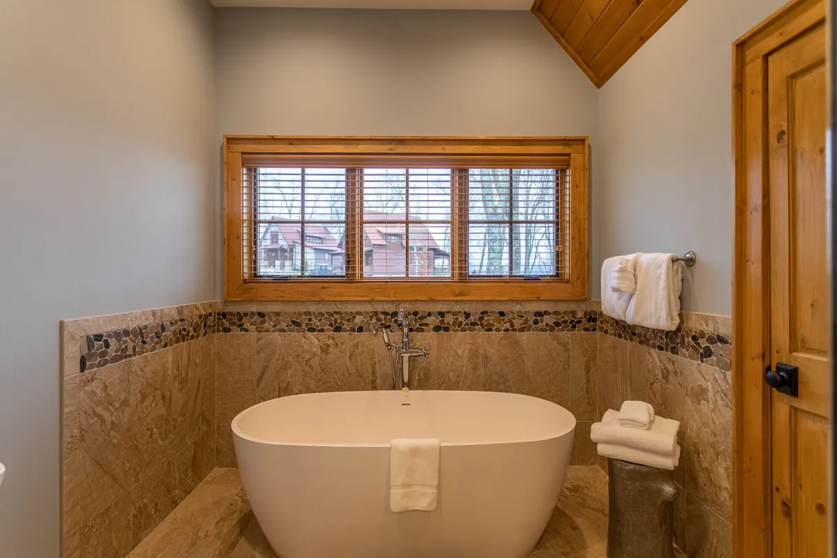 A tranquil, spa-inspired bathroom with a luxurious freestanding soaking tub set beside a wooden window, textured wall tiles, and a serene mood. This space is ideal for relaxation and self-care, echoing the spa-like qualities outlined in the master suite.