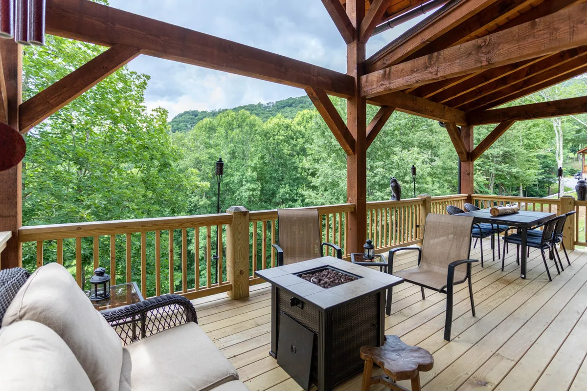 A wooden porch with sturdy beams features a gas fire pit table surrounded by plush, comfortable seating. Lush greenery provides a serene backdrop, and the space is bathed in the warm light of either morning or evening, evoking a sense of relaxation and connection with nature.