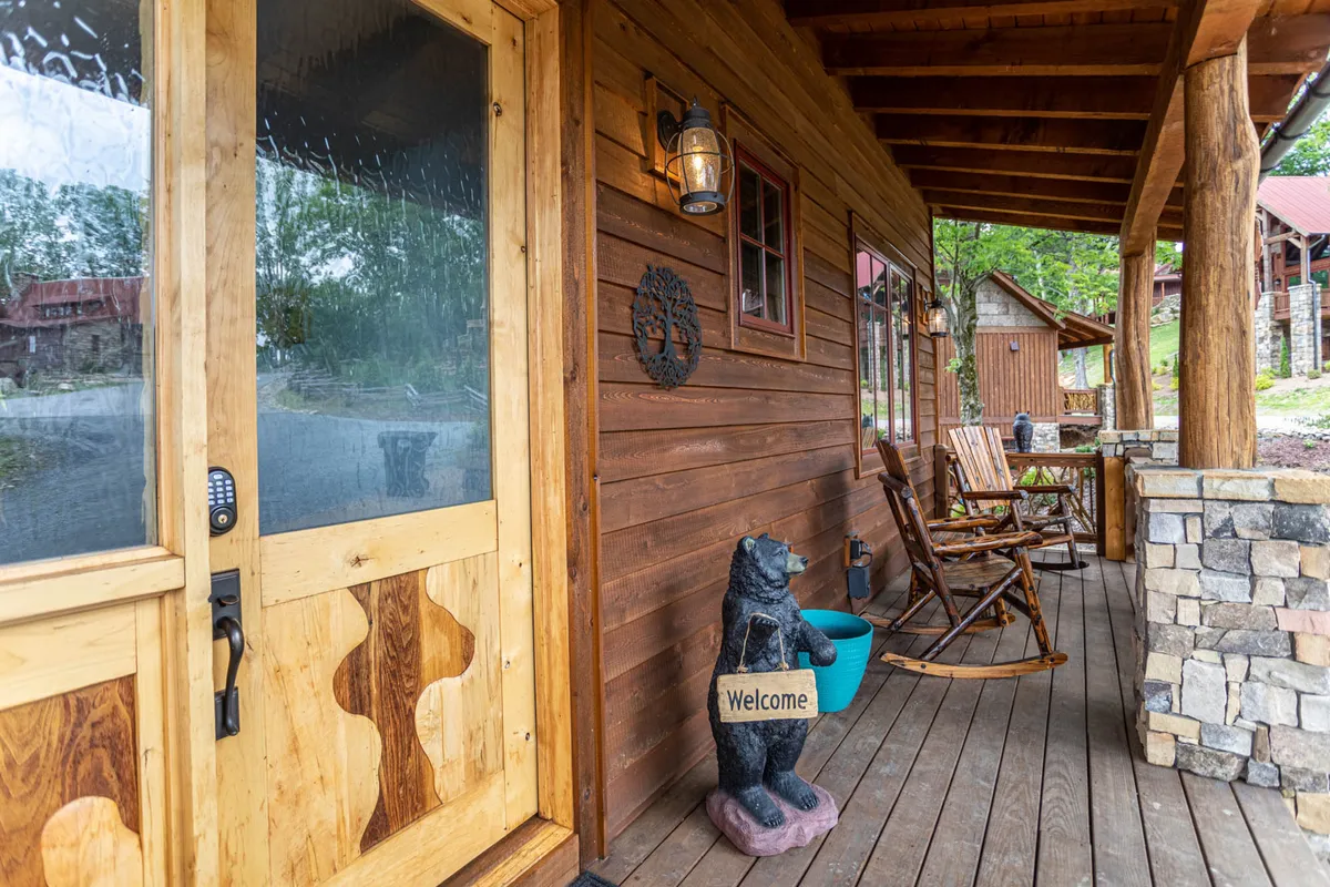 This image captures the exterior entrance of Bag End, featuring a welcoming wooden door with an animal motif, a whimsical bear statue, classic rocking chairs, and a sturdy stone column. The porch setting offers a view of the tranquil natural surroundings.