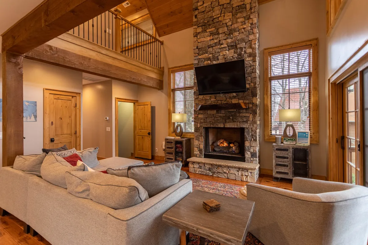 A spacious living room with a grand stone fireplace as its focal point, surrounded by large windows that offer views of the outdoors. Comfortable sofas are arranged for conversation or relaxing with a book. The cozy ambiance is enhanced by the warm glow of the fire, perfect for winter nights or relaxing after outdoor adventures.