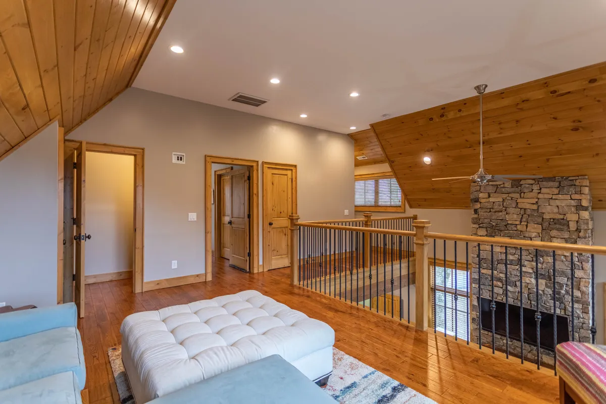 A spacious, stylish living area with a grand stone fireplace, wooden ceiling, balcony railing, and plush seating. Large windows suggest expansive views, and the balcony hints at opportunities for socializing while enjoying the outdoors.